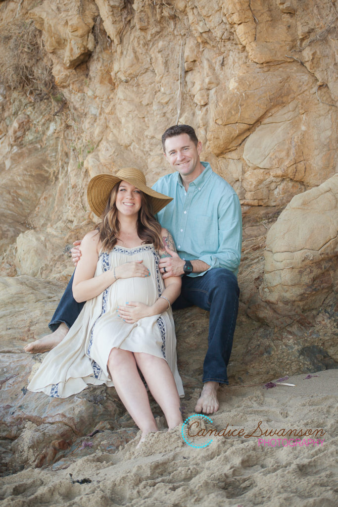 Expecting mom, holding bump, with husband behind her sitting on a rock. 
