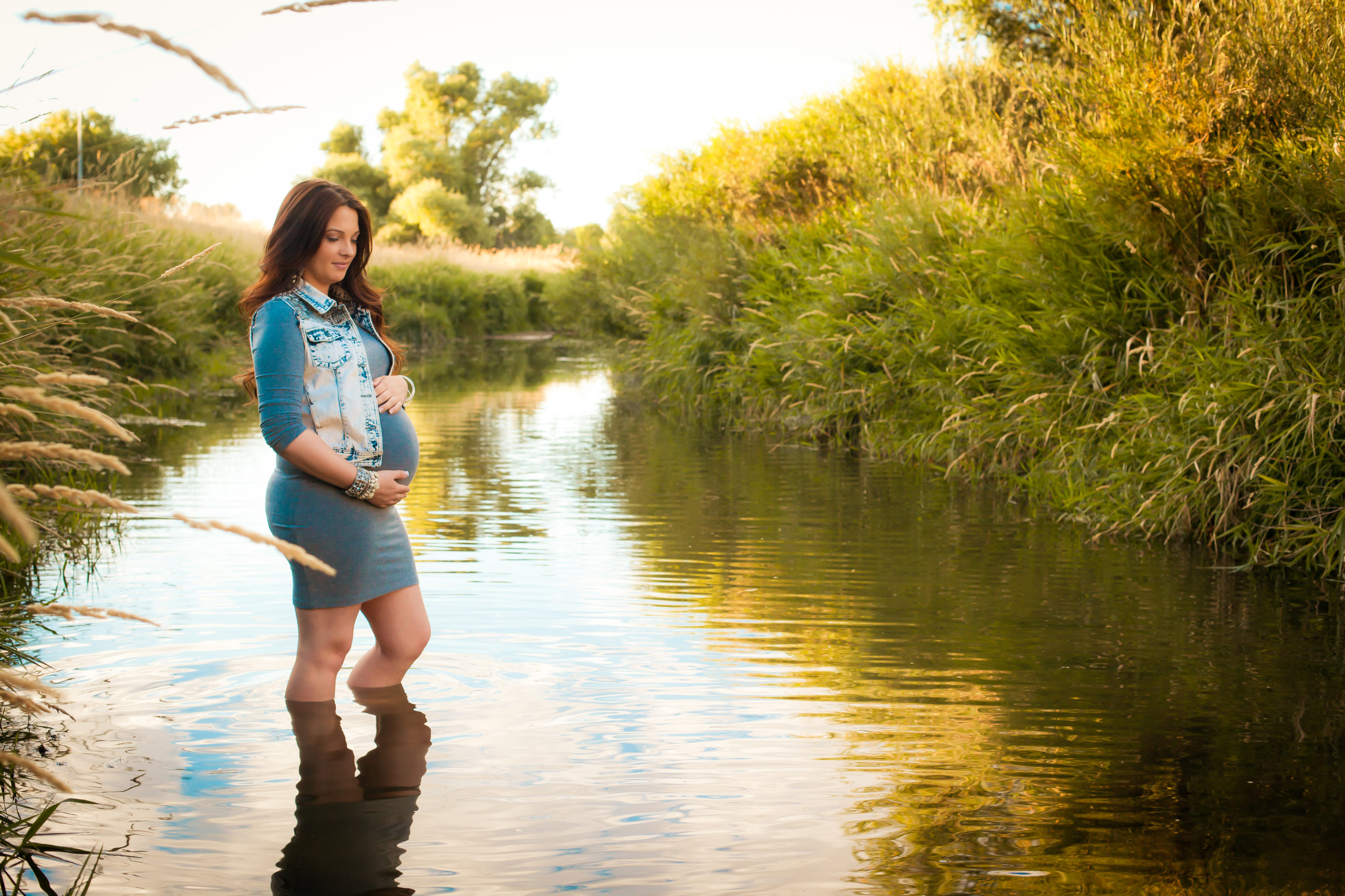 Pregnant mom, posing sideways while holding bump, standing in a river during golden hour.