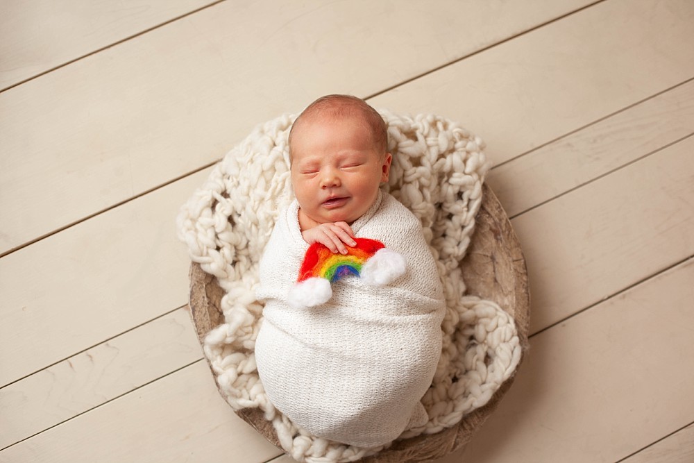 Rainbow baby sitting in wooden bowl, wrapped in cream blanket holding small felt rainbow.